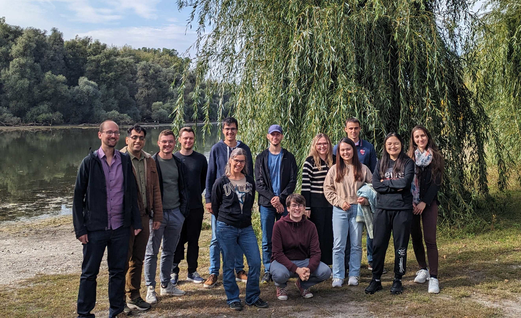 Photo of the AiMat group in October 2023 standing by a lake with trees in the background.